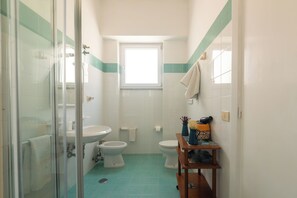 A bathroom with a glass shower enclosure, a sink, a toilet, and a bidet. There is a window above the toilet, and a small wooden shelf with decorative items. - Villa dei Sogni (Ischia)