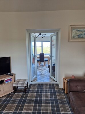 A living room with a television, a sofa, a dining area with chairs, and a view through a glass door.