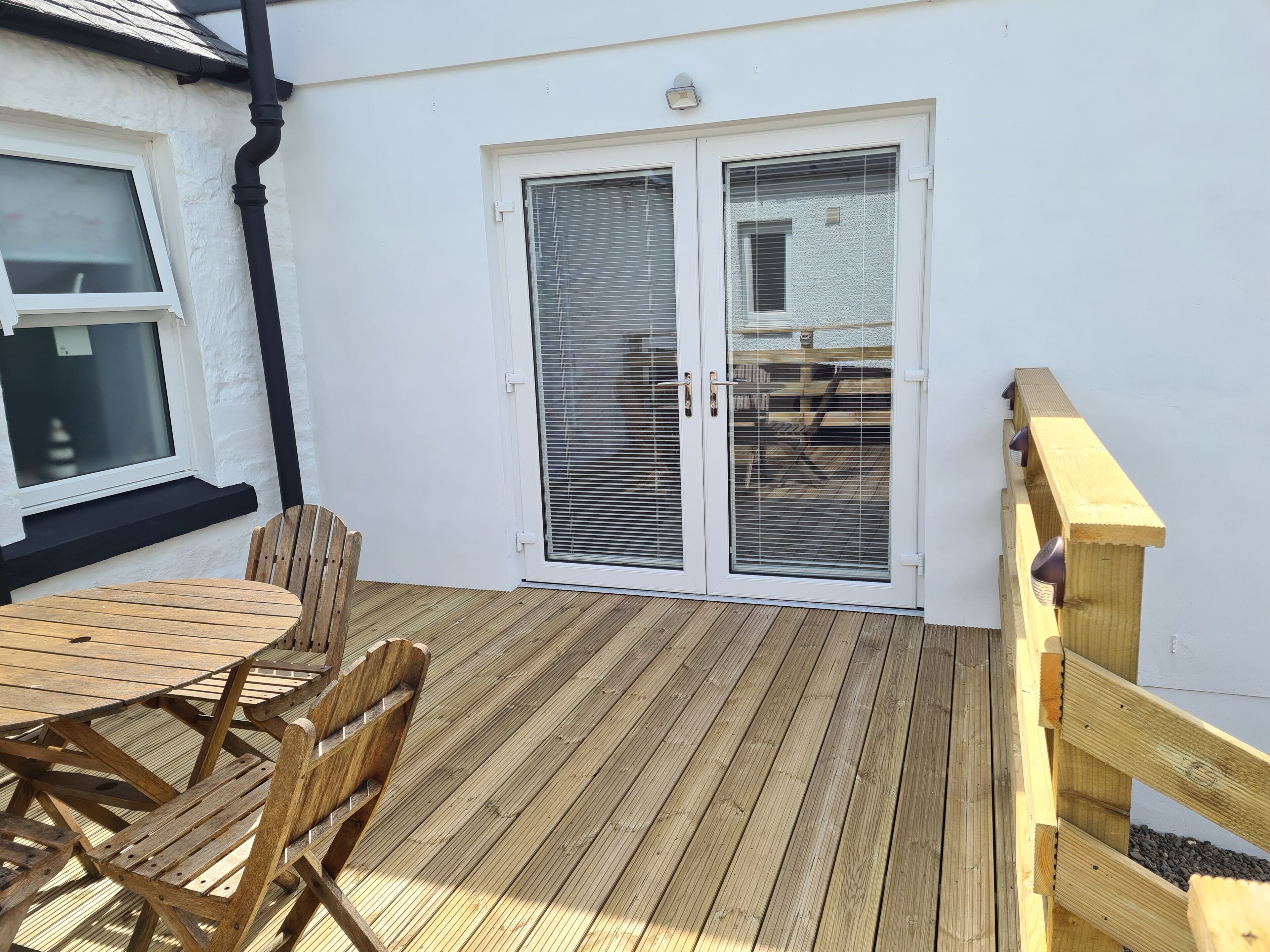 Une terrasse en bois avec une table et des chaises, une porte-fenêtre coulissante et un mur extérieur blanc.
