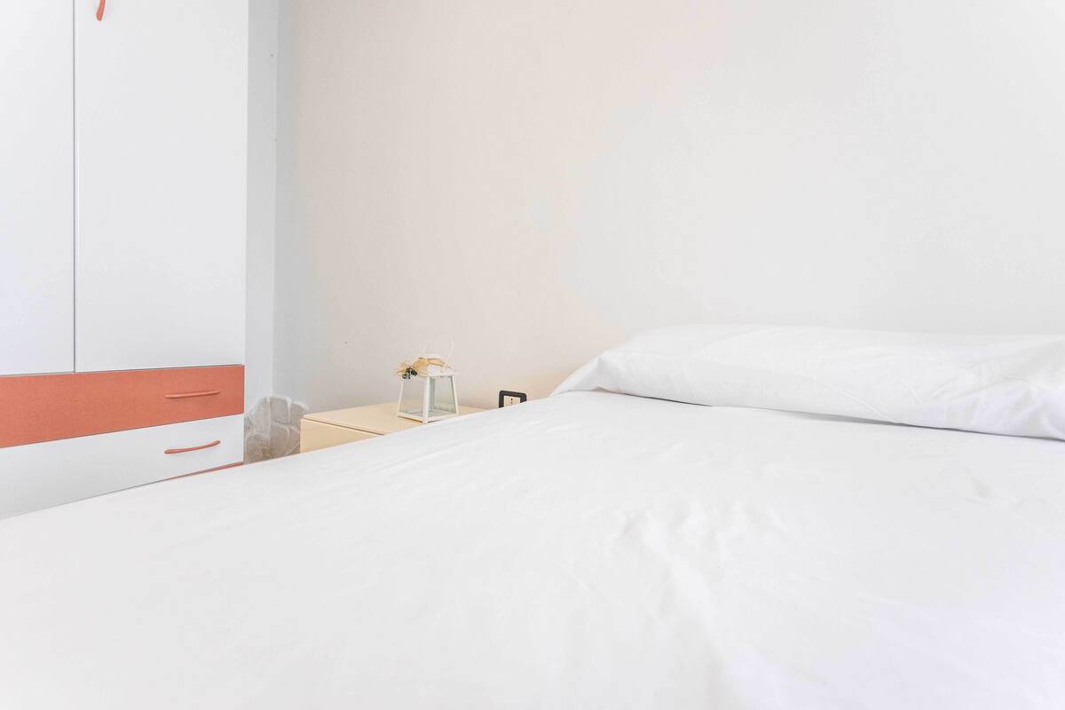 Cama bem arrumada com lençóis brancos, mesa de cabeceira com abajur e guarda-roupa com detalhe vermelho.