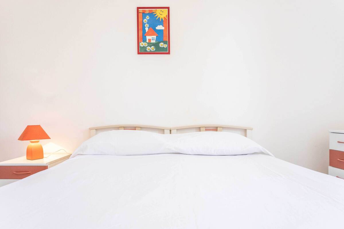 Uma cama bem arrumada com lençóis brancos, cabeceira de madeira, mesa de cabeceira com abajur e um quadro na parede.