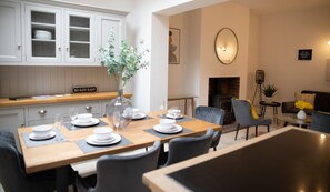 Un coin repas avec une table en bois dressée pour quatre personnes, un vase avec une plante, et une cuisine avec des armoires blanches et un panneau sur lequel on peut lire : « DÉBITANTS INTERDITS ».