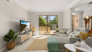 A modern living room with a flat-screen TV, a grey sofa, a white coffee table, and a large sliding glass door leading to a balcony.