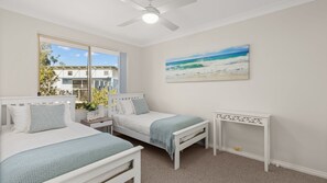 Een slaapkamer met twee bedden, een plafondventilator, een schilderij van een strand en uitzicht op een gebouw.