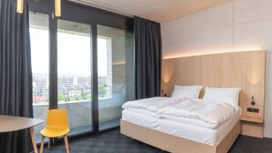 Een moderne hotelkamer met een groot bed, een klein tafeltje, een gele stoel en uitzicht op de stad door grote ramen.