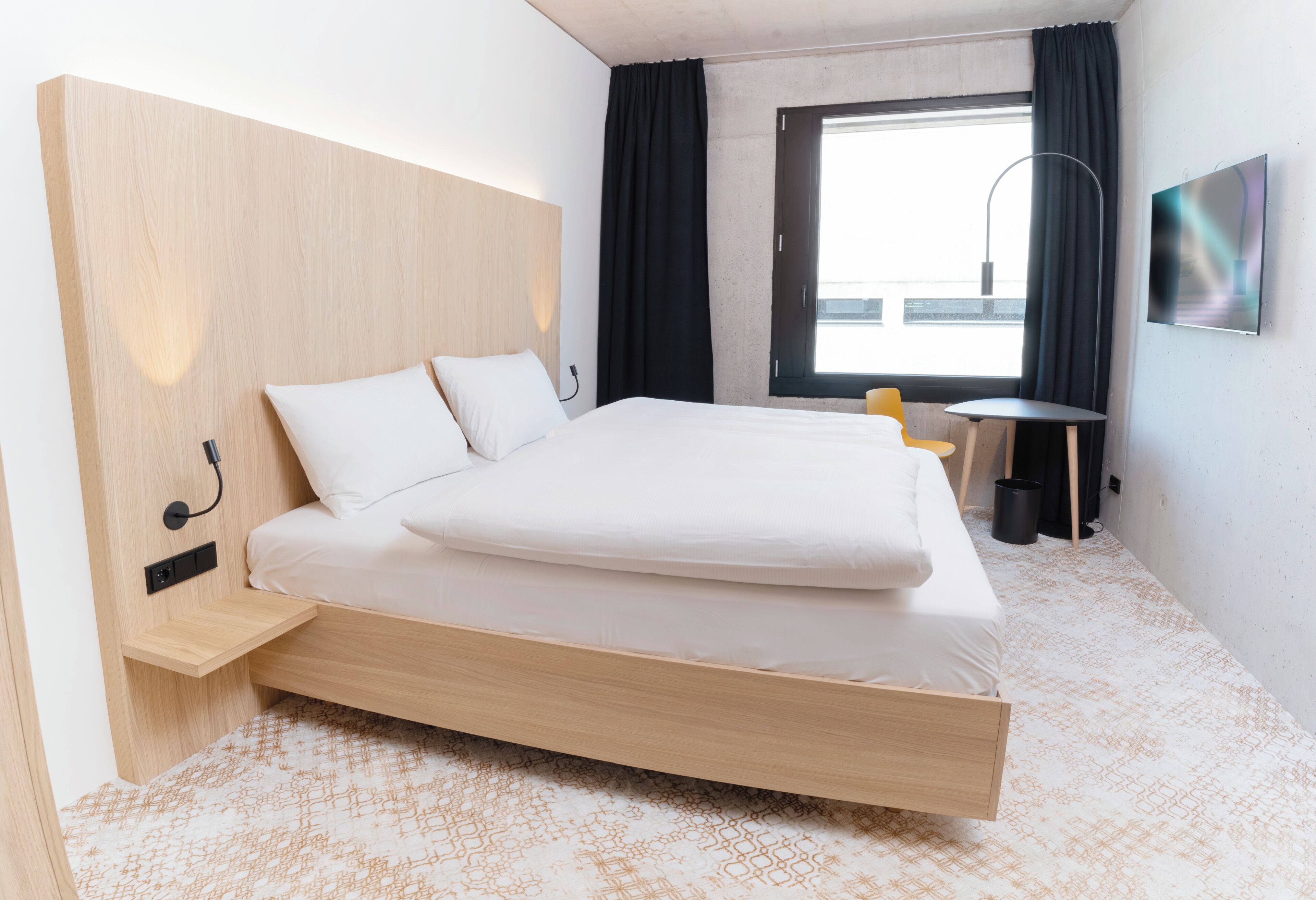 A modern hotel room with a wooden bed, a small table, a chair, a TV, and a window with curtains.