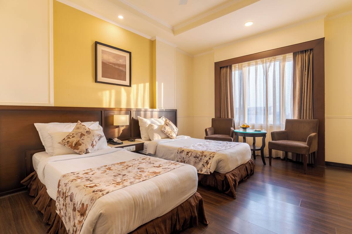 A hotel room with two beds, a sitting area with a chair and a small table, a framed picture on the wall, and a window with curtains.