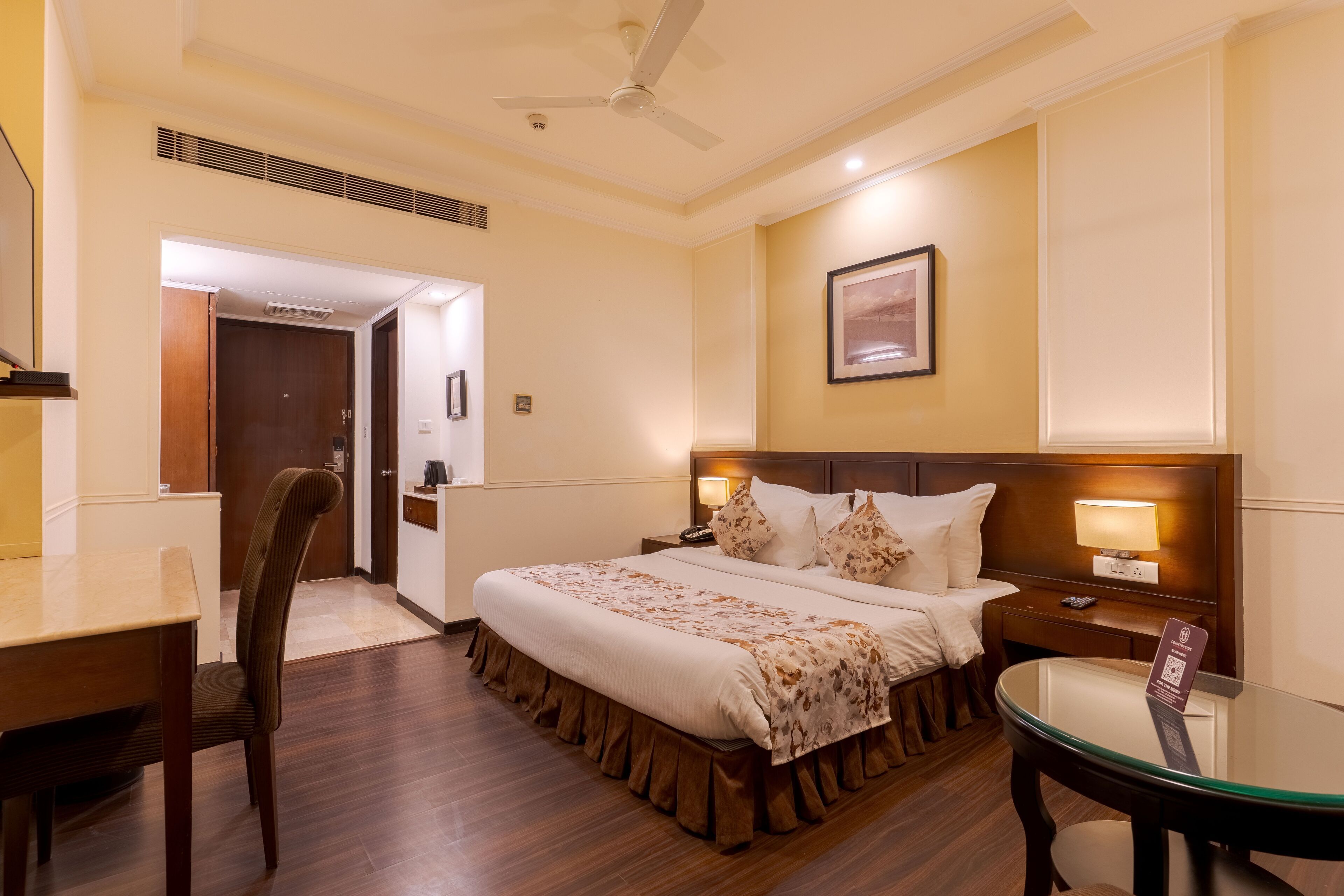 A hotel room with a bed, a desk, a chair, a nightstand, and a glass table.