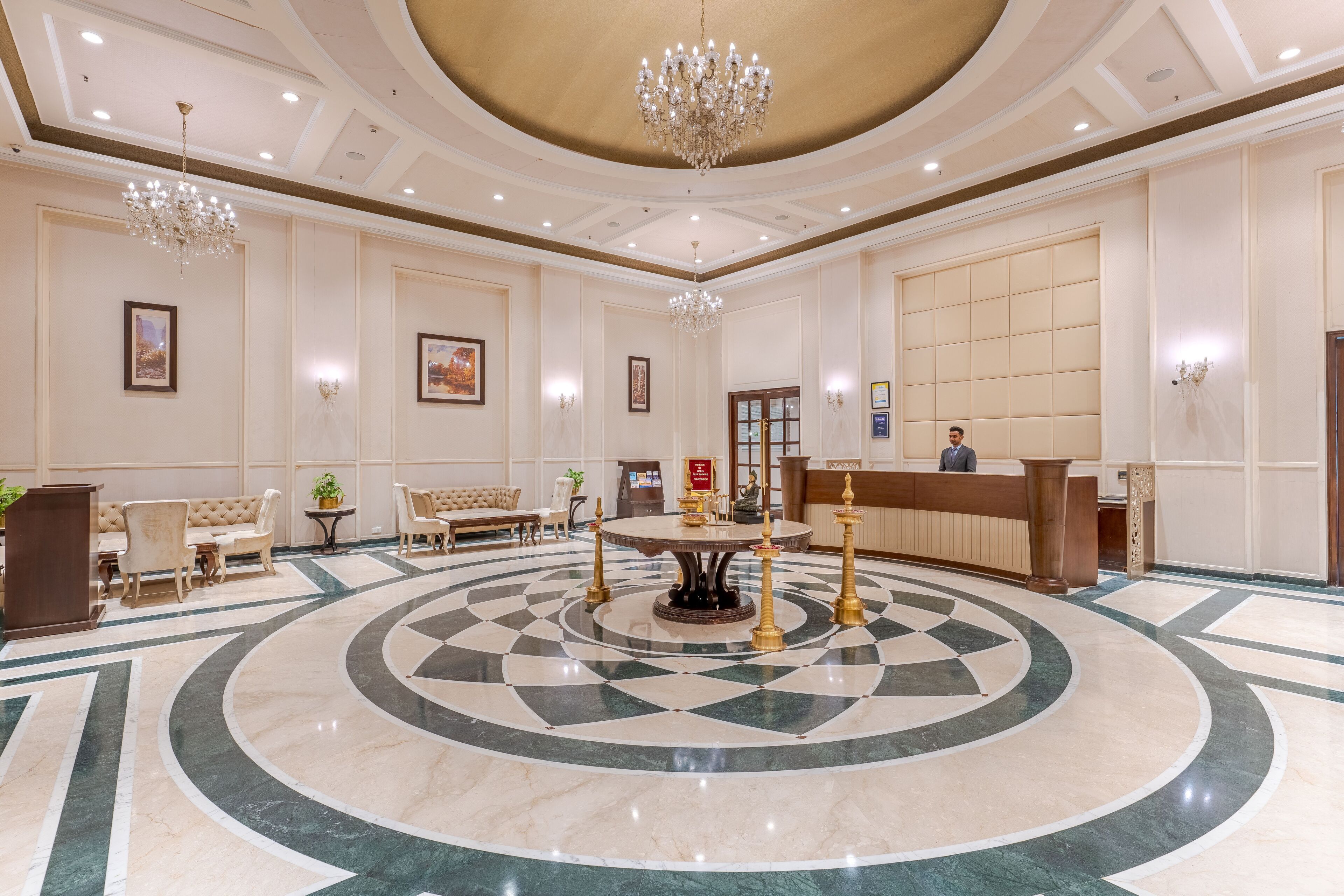 A lobby with a central round table, sofas, a reception desk, and chandeliers.