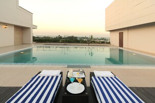 Área da piscina com duas espreguiçadeiras, uma mesa com bebidas e um pequeno tabuleiro, e vista para o horizonte urbano ao fundo.