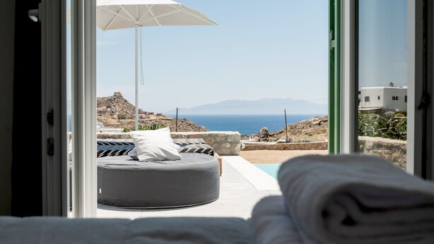 Un dormitorio con vista al mar, una cama, un paraguas blanco y una pequeña zona de estar circular.