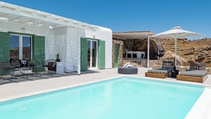 Un área de piscina con una pérgola blanca, contraventanas verdes, mobiliario de exterior y un muro de piedra de fondo.