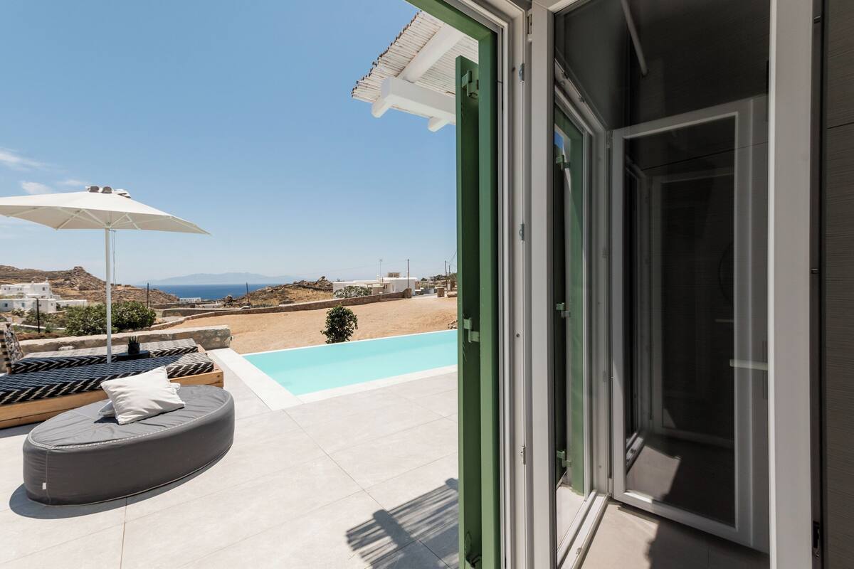 A modern outdoor seating area with a pool, a white umbrella, and a view of the sea and hills.