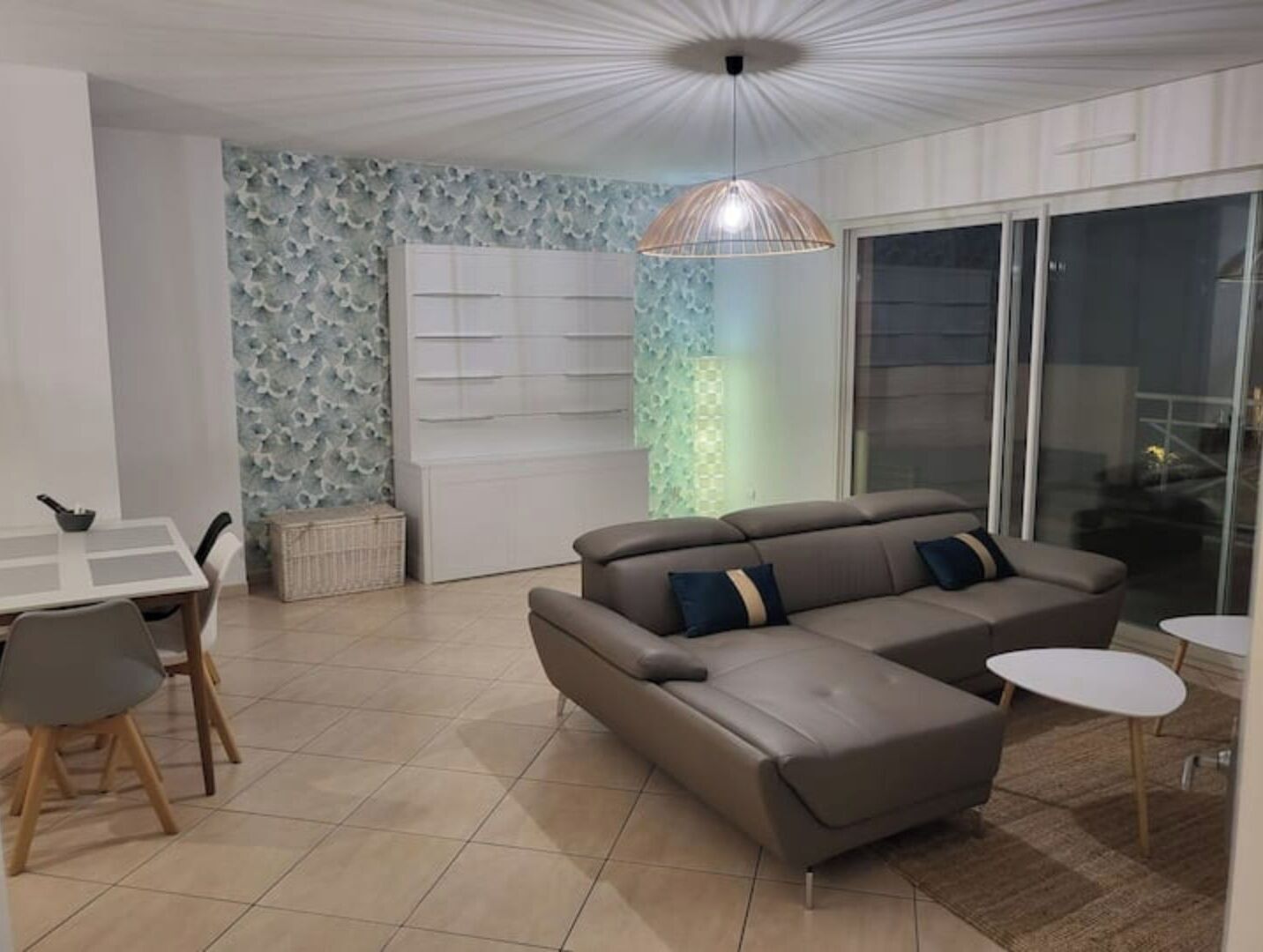 A modern living room with a sectional sofa, a dining table with chairs, a decorative wall, and a large window.