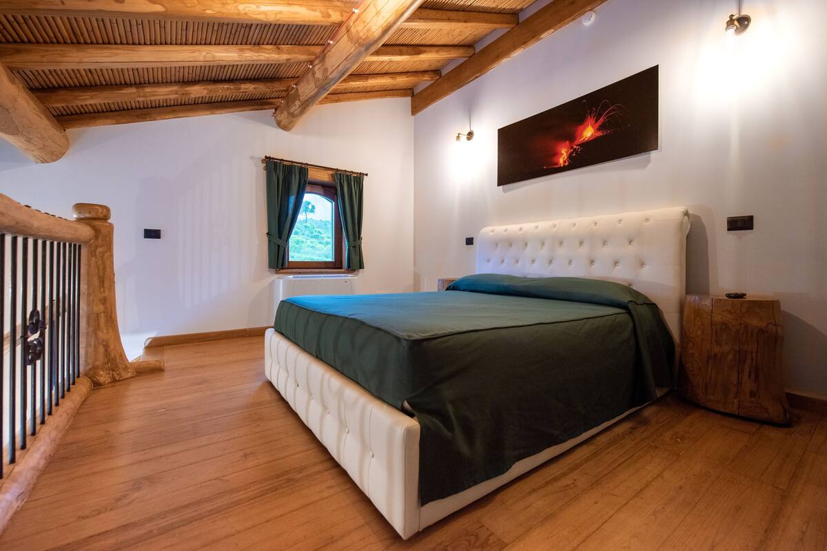 A bedroom with a wooden ceiling, a bed with a green bedspread, a window with curtains, and a wall-mounted artwork.