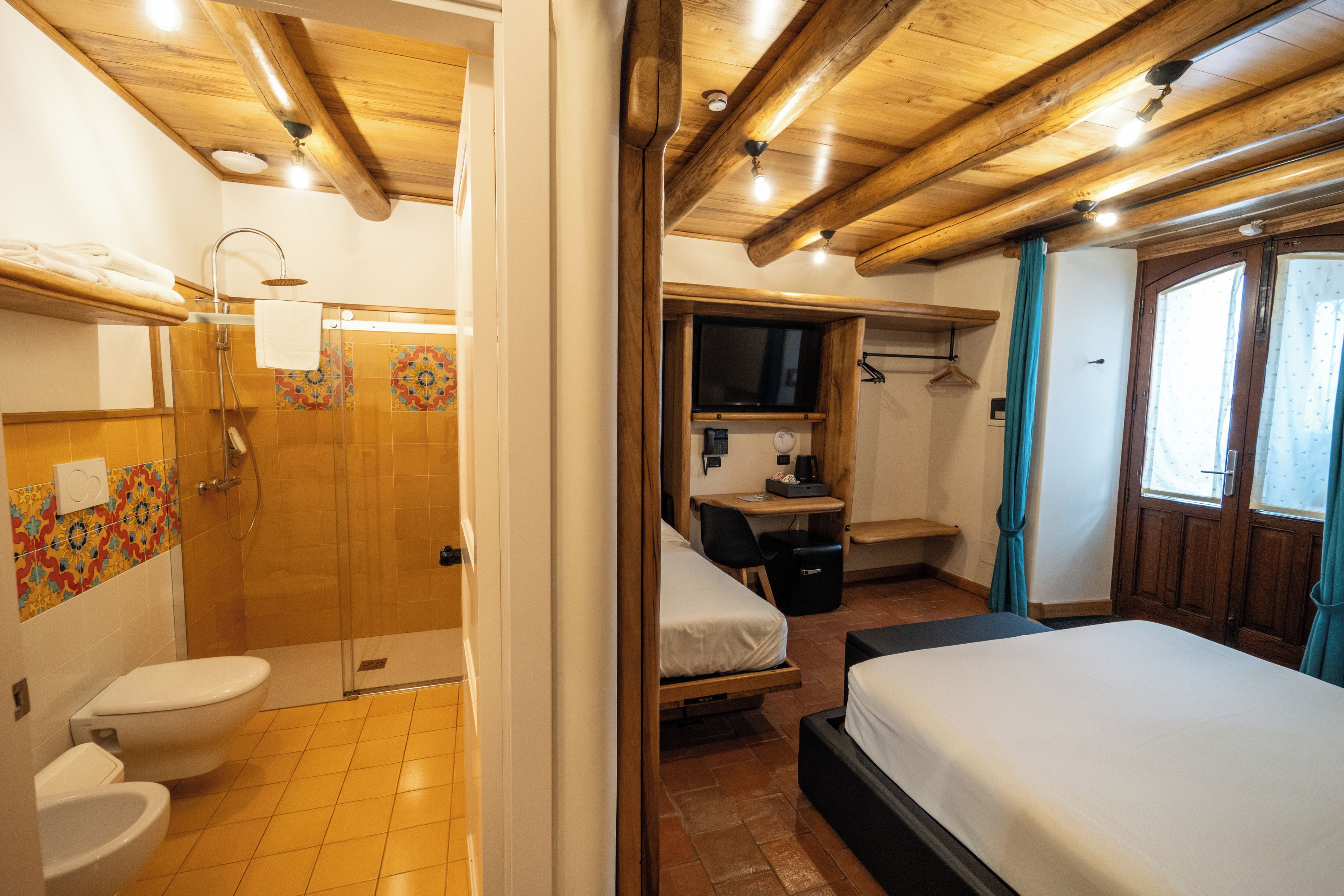 A room with a wooden ceiling, a bed, a shower, and a small desk with a television.
