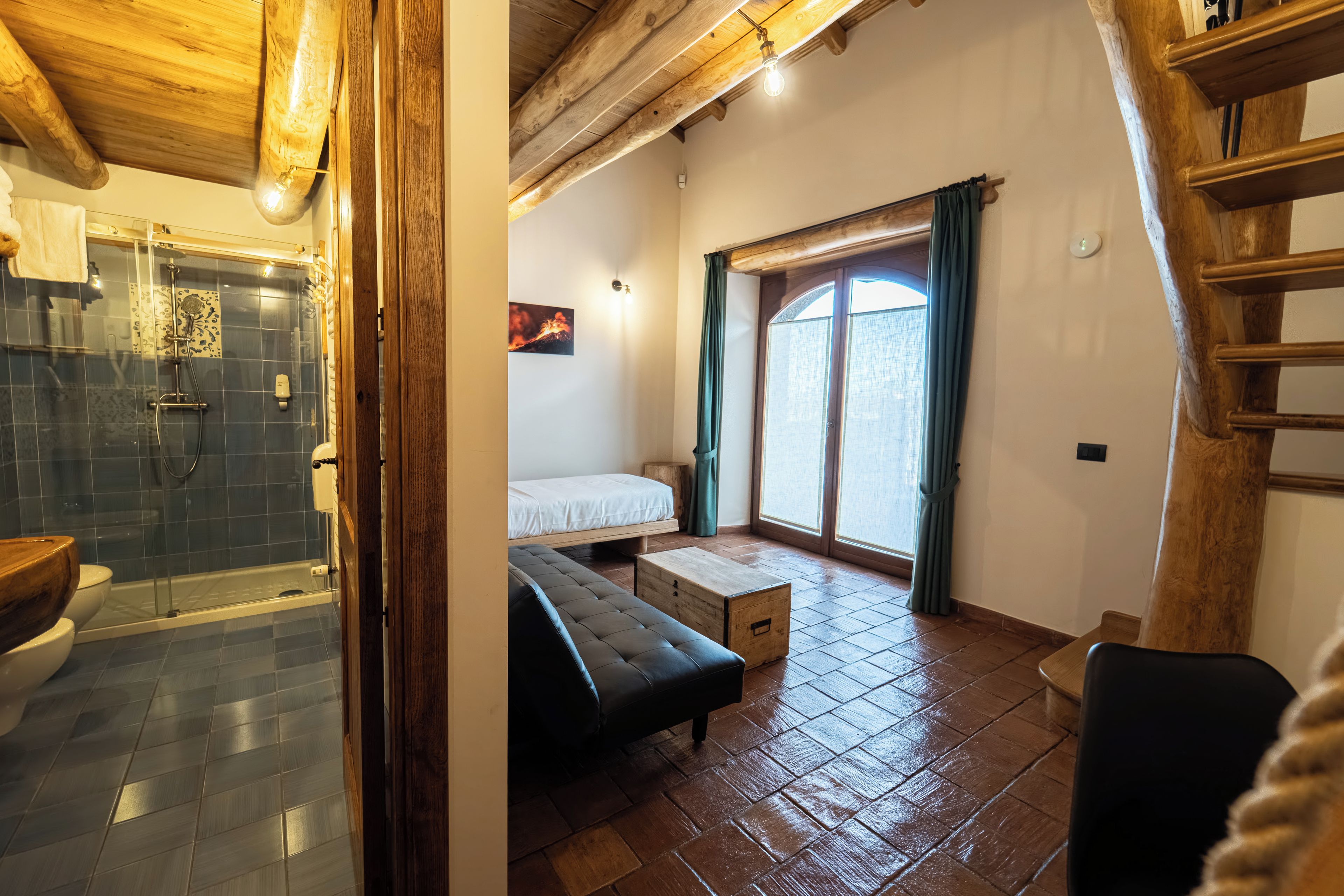 A room with a shower, a bed, a bench, and a window with curtains.