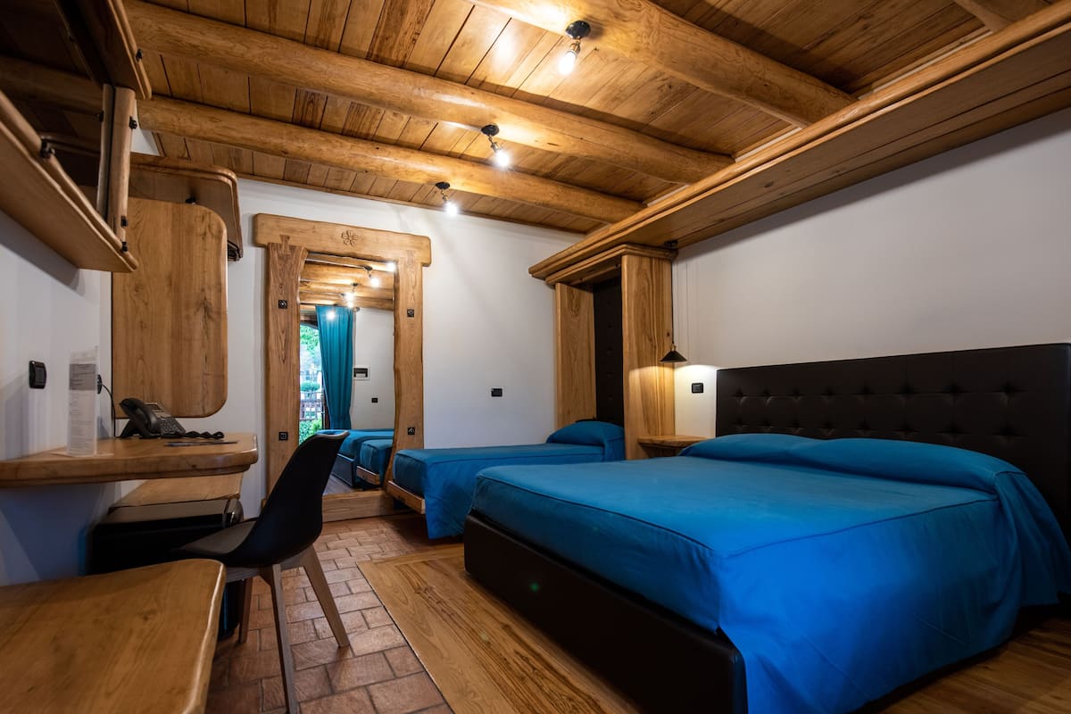 A room with wooden ceiling beams, a bed with blue bedding, a desk with a telephone, and a chair.