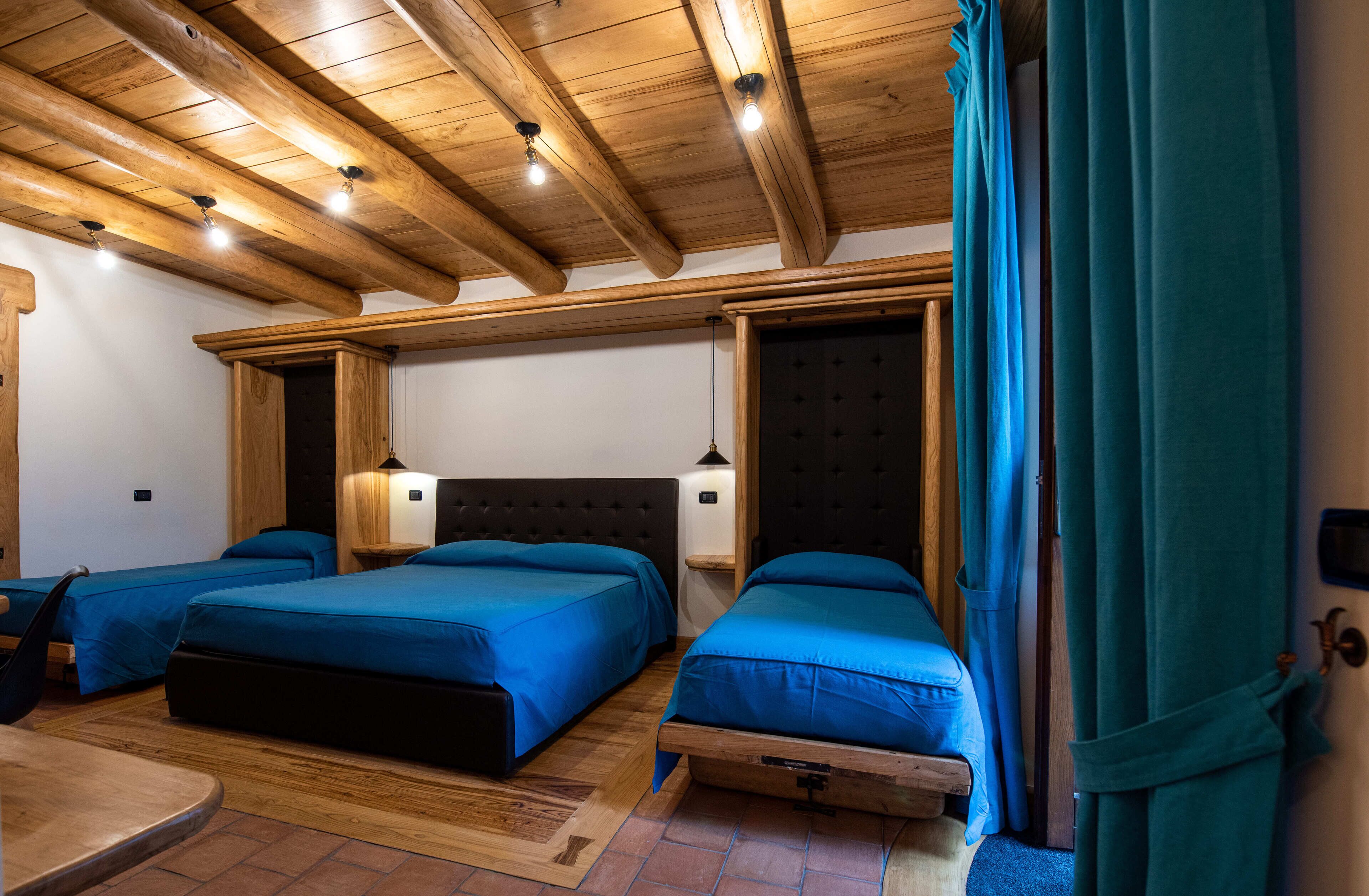 Een kamer met houten balken in het plafond, drie bedden met blauw beddengoed, een houten tafel en een deur met turquoise gordijnen.