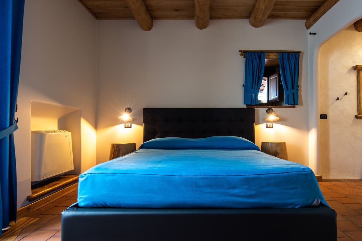 A bedroom with a bed, two bedside lamps, a window with curtains, and a wooden ceiling.