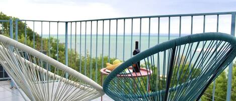 A balcony with two chairs, a table with a bottle and fruit, and a view of the sea.