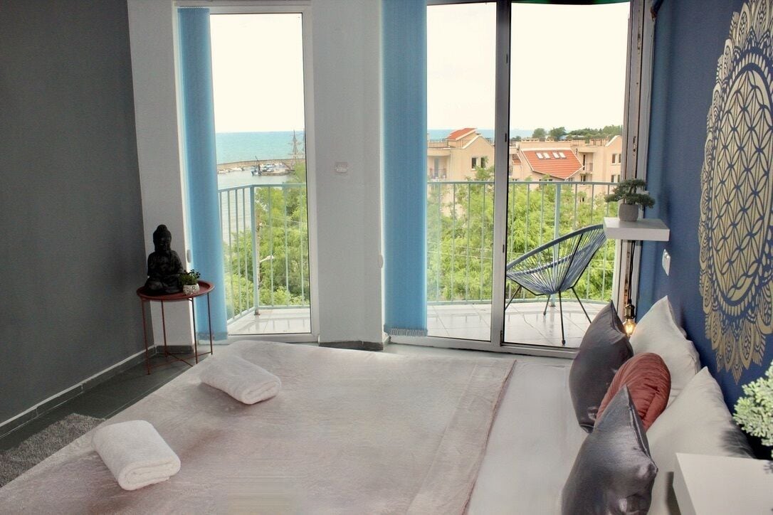 Une chambre moderne dotée d’une grande porte-fenêtre coulissante donnant sur un balcon avec vue sur la mer et les bâtiments.