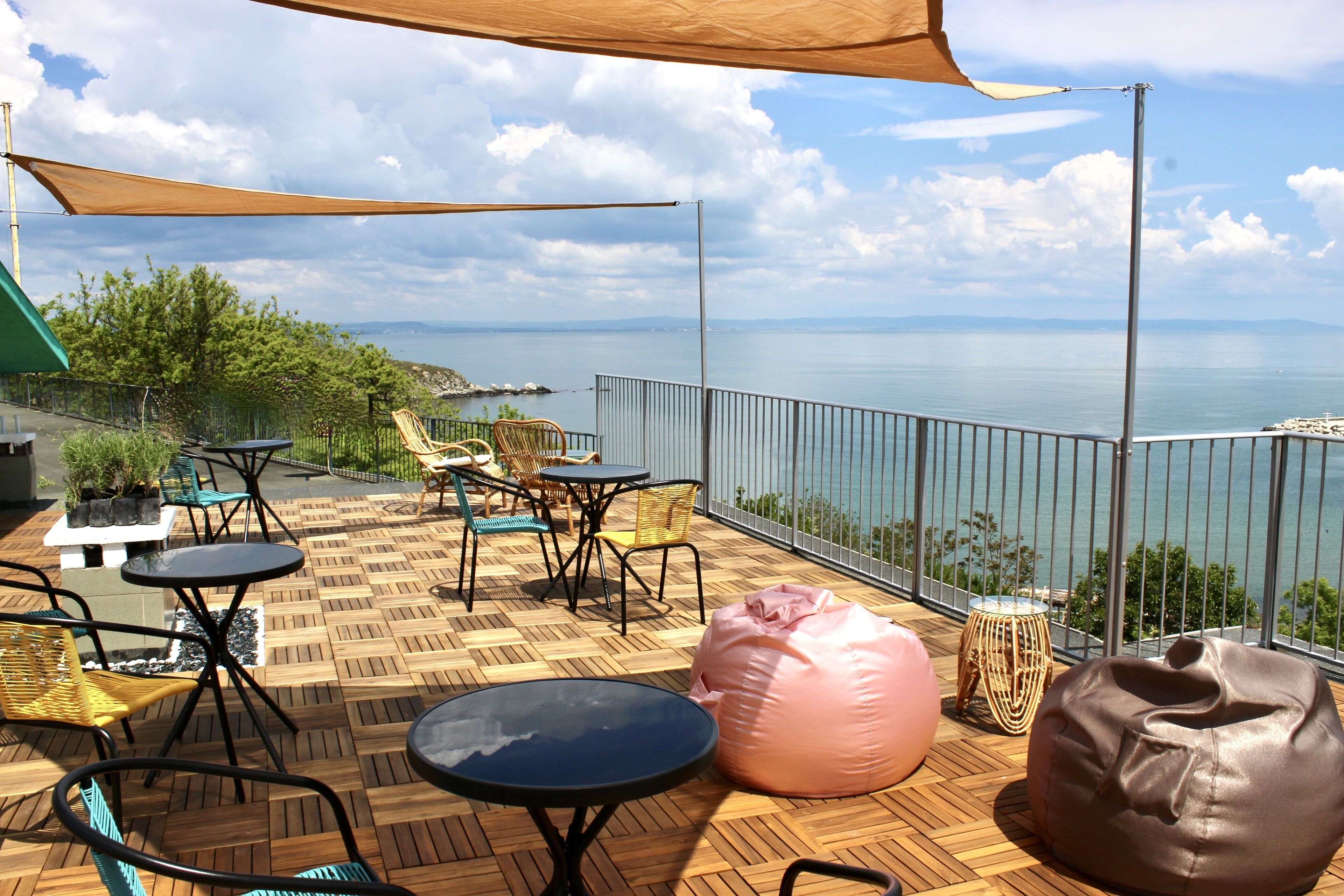 An outdoor terrace with wooden flooring, metal tables and chairs, a large bean bag, and a view of the sea.