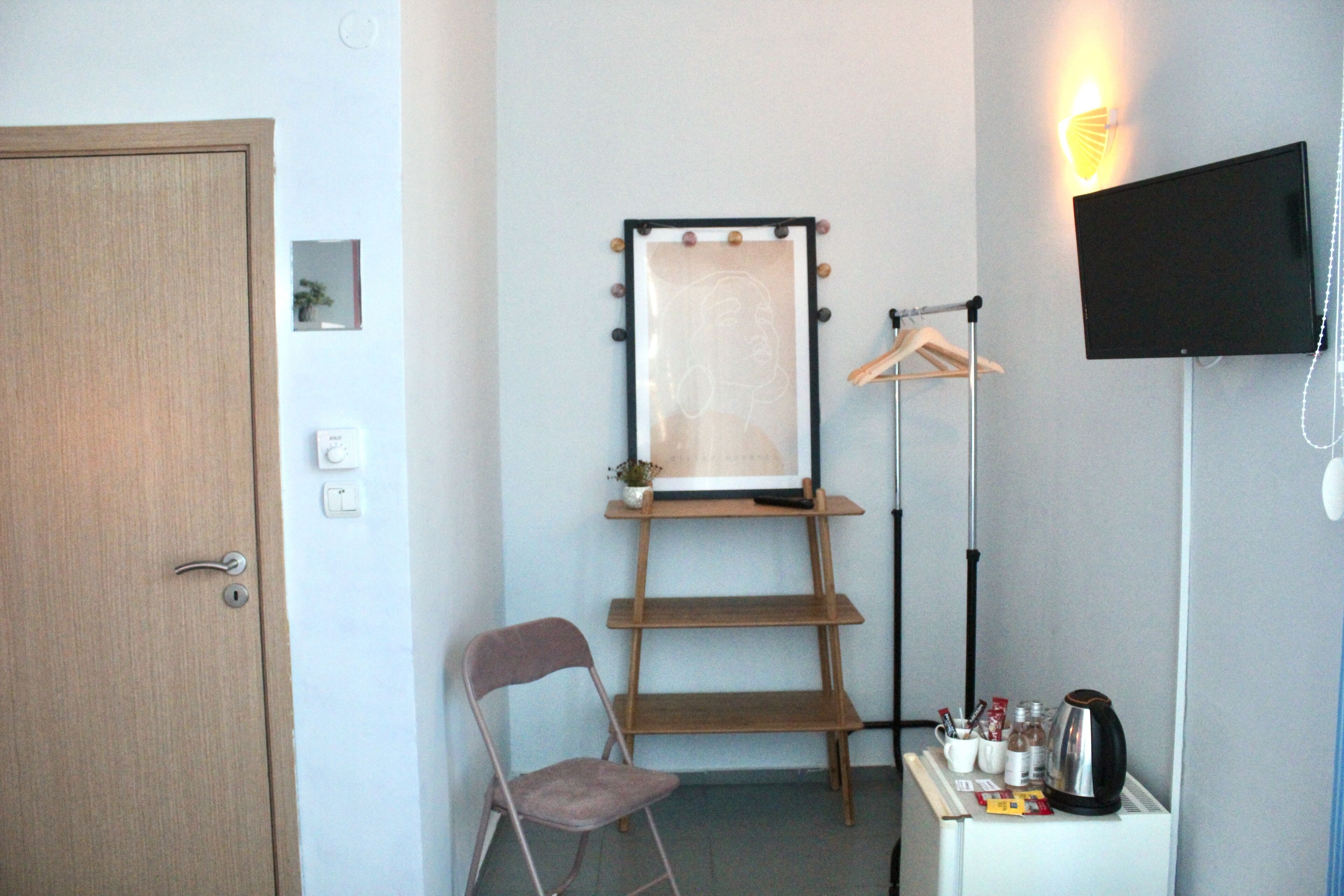Een kamer met een houten deur, een stoel, een klein tafeltje met een waterkoker, een ingelijst beeld op een standaard, en een televisie aan de muur.
