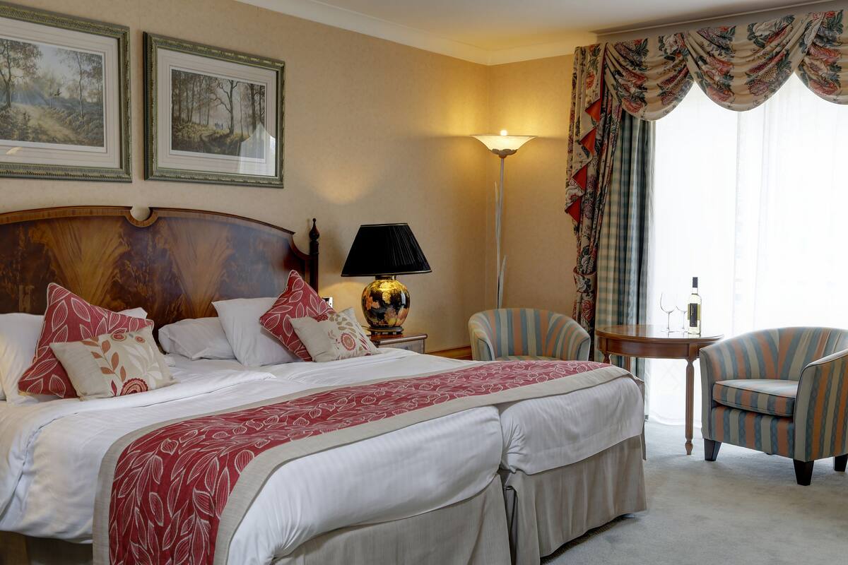 A hotel room with a large bed, two armchairs, a small table with a lamp, and a view of the outdoors through a window with curtains.