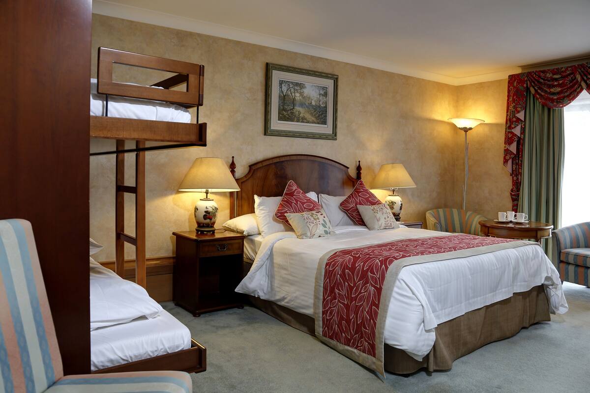 A hotel room with a bunk bed, two bedside tables with lamps, a chair, a desk with a cup, and a framed picture on the wall.