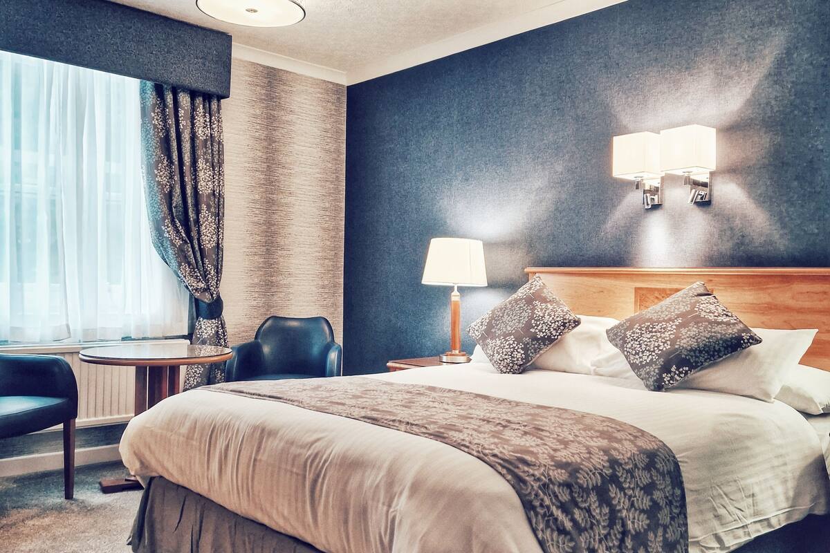 A hotel room with a large bed, two bedside lamps, a headboard, a round table, a chair, and a window with curtains.