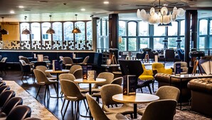 A modern lounge area with multiple seating options including round tables, sofas, and chairs. Large windows provide natural light and views of the outdoors. - Blunsdon House Hotel (Swindon)
