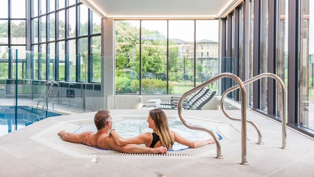A couple relaxing in a hot tub with a view of a swimming pool and greenery outside. - Blunsdon House Hotel (Swindon)
