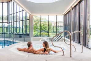 A couple relaxing in a hot tub with a view of a swimming pool and greenery outside. - Blunsdon House Hotel (Swindon)