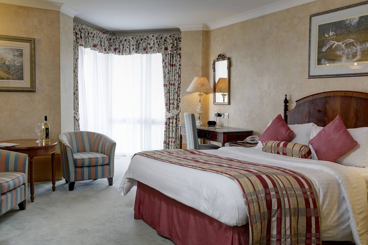 A hotel room with a large bed, a desk with a lamp, a chair, and a window with curtains.