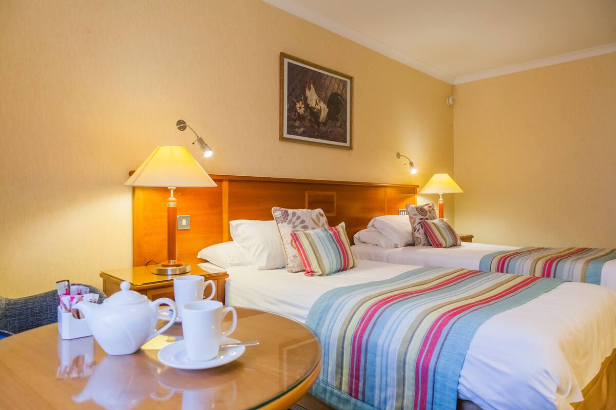 A hotel room with two beds, a wooden table with a teapot and cups, and two lamps.