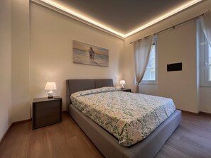 A bedroom with a large bed, two bedside tables with lamps, a painting on the wall, and a window with curtains. - Val Di Sole (Rapallo)