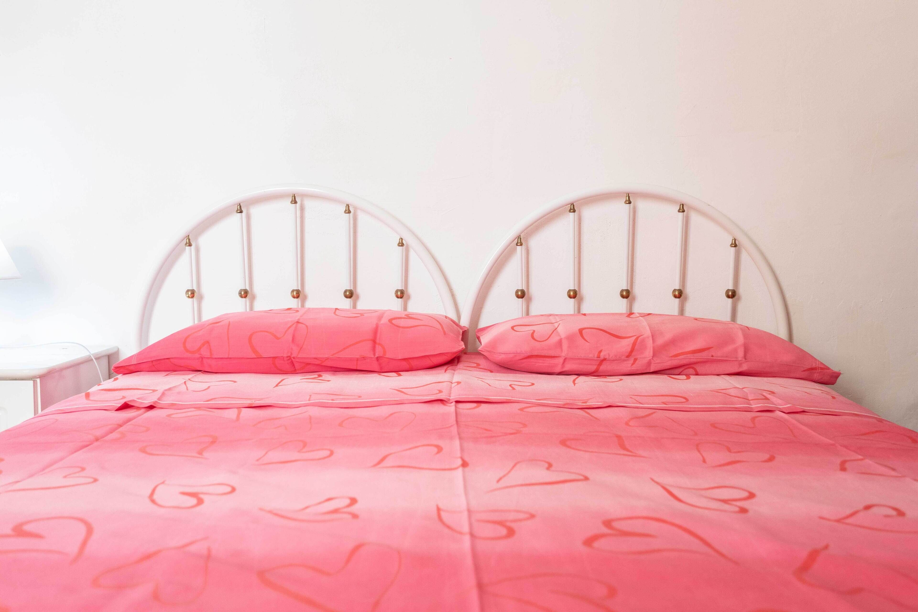 Cama com estrutura de metal branco e roupa de cama rosa com estampas de corações.