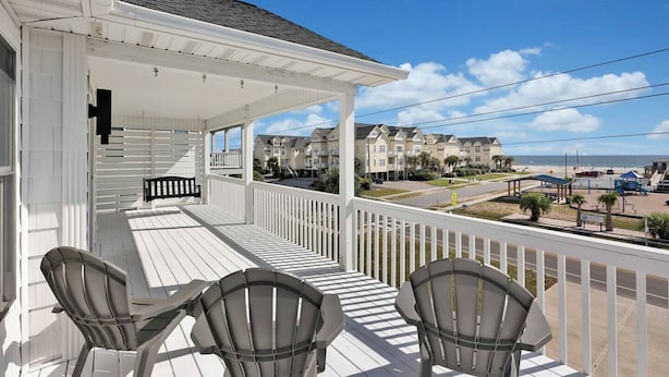 A covered porch with two Adirondack chairs, a view of a beachside community, and a clear sky.