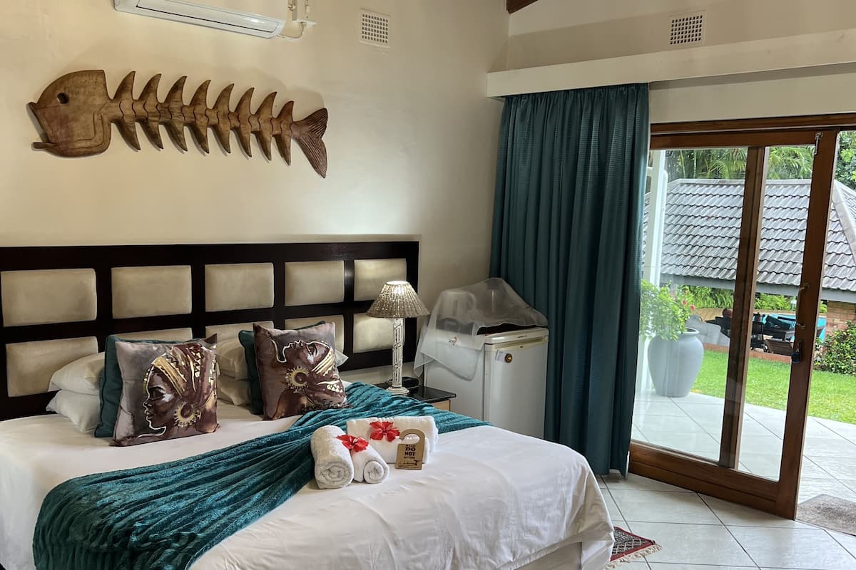 Quarto com cama grande, decoração de peixe de madeira na parede, ventilador de teto e vista para o exterior por uma porta de vidro.