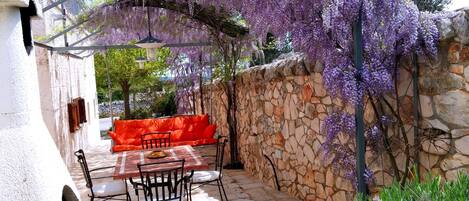 A stone-walled outdoor seating area with a table and chairs, under a pergola covered with purple wisteria flowers.