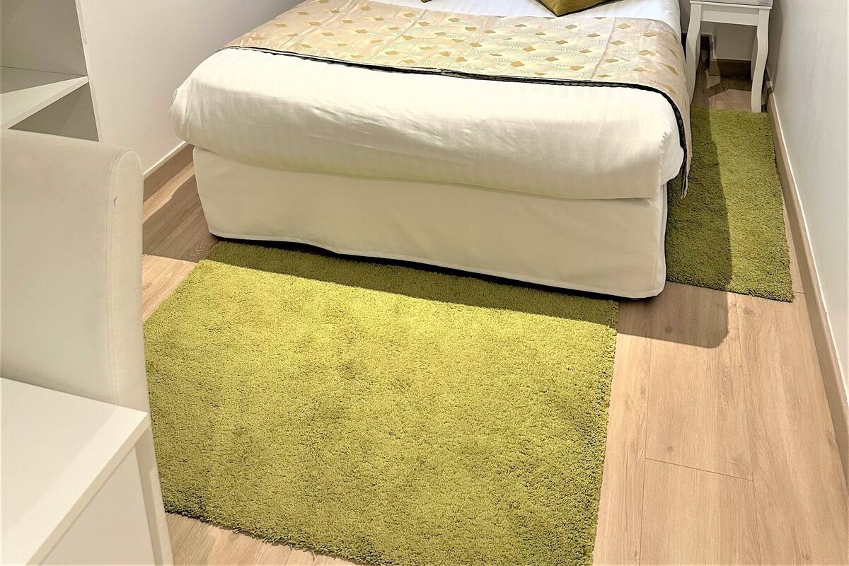 A bedroom with a bed, two pillows, a nightstand, a window with curtains, and a green rug.