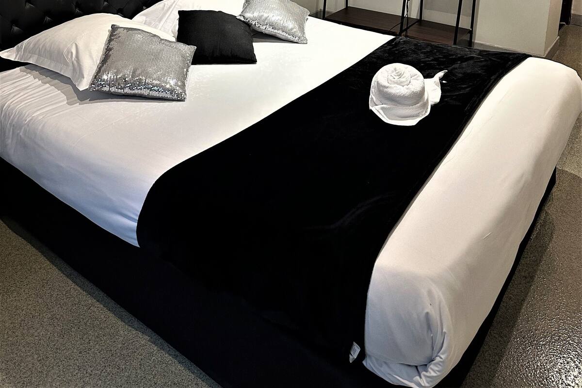 A neatly made bed with a black headboard, white and silver pillows, a black and white throw blanket, and a white towel on the foot of the bed.