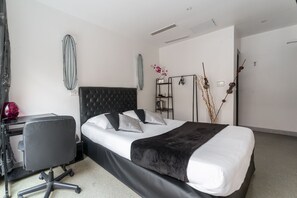 Een moderne hotelkamer met een zwart bekleed hoofdeinde, een groot bed met witte lakens, een zwart bureau met stoel en een klein nachtkastje.