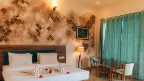 A hotel room with a large bed, wooden headboard, a wall-mounted picture, a green curtain, and a wooden chair. - The Sky Imperial Pavoreal Jungle Resort (Kumbhalgarh)
