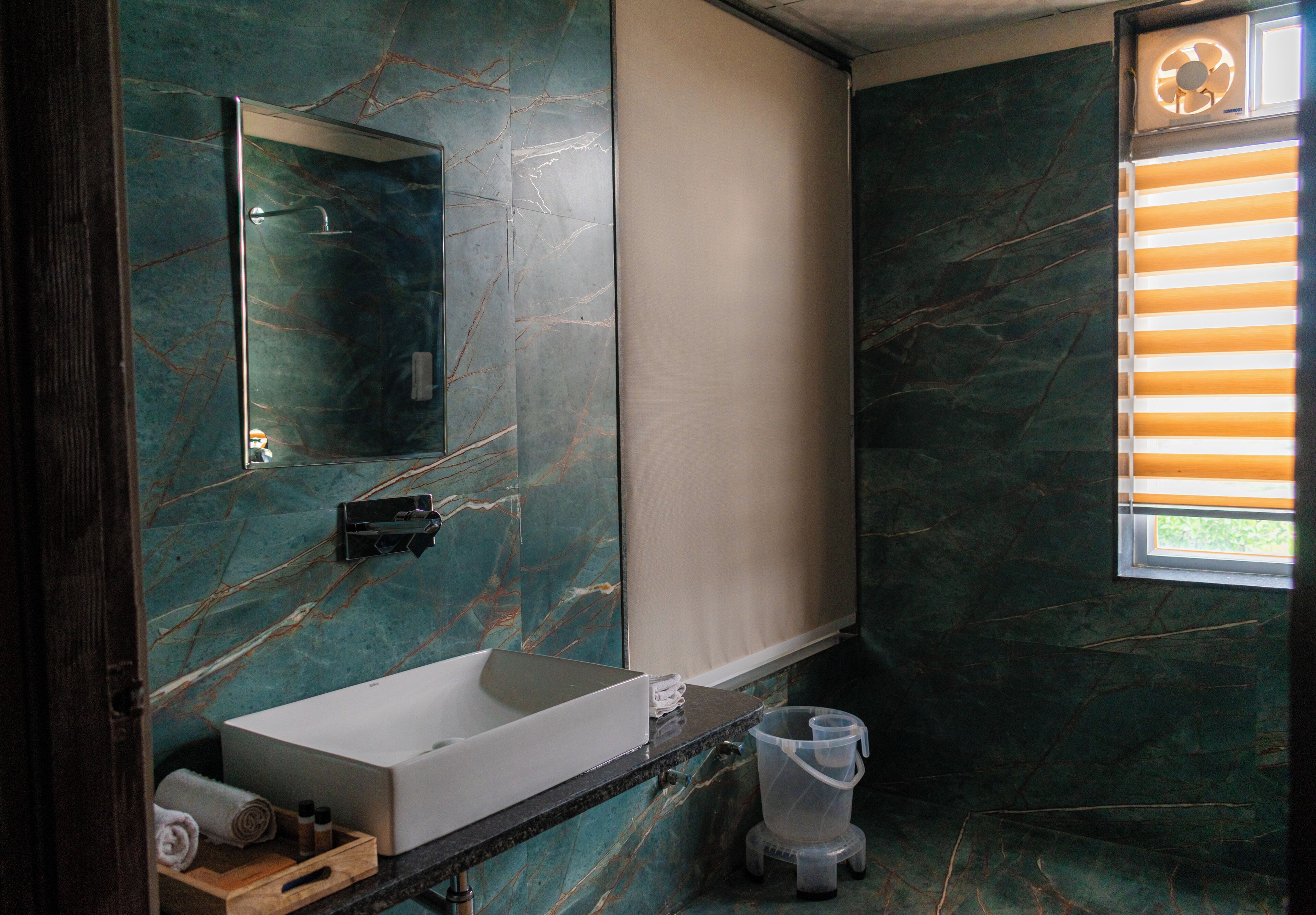 A bathroom with a marble wall, a white sink, a mirror, and a window with blinds.