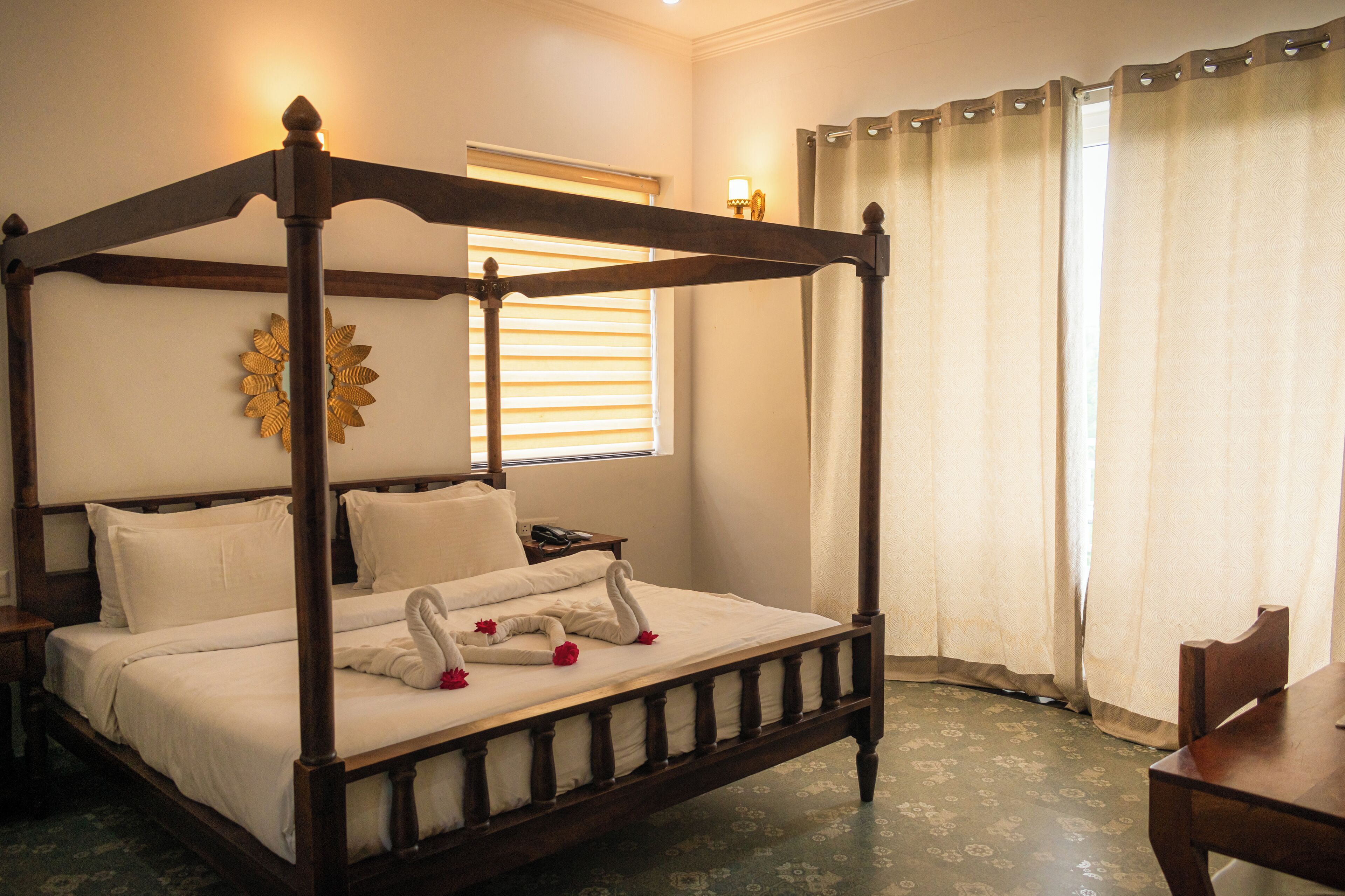 A four-poster bed with white linens and decorative swans, a bedside table with a phone, and a window with blinds.