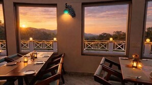 A restaurant with tables and chairs, large windows with views of a sunset and hills, and string lights on the balcony railing. - The Sky Imperial Pavoreal Jungle Resort (Kumbhalgarh)