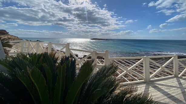 Vista de platja amb una tanca blanca, palmeres i un cel serè.