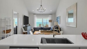 A modern kitchen with a central island, a dining area with a sofa and chairs, and a view of the sea through large windows.